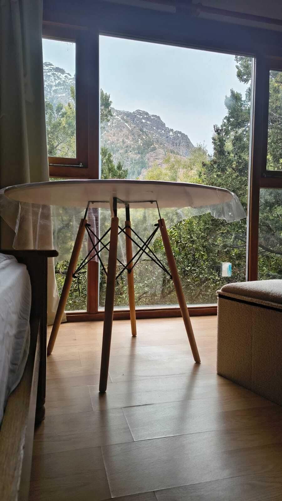 Une pièce dotée d’une grande fenêtre, d’un plancher en bois, d’un lit, d’une table sur laquelle est posée une planche de surf, et qui offre une vue sur un paysage montagneux.