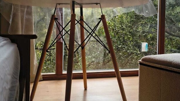 Ein Zimmer mit großem Fenster, Holzboden, einem Bett, einem Tisch mit Surfbrett und Blick auf eine gebirgige Landschaft.