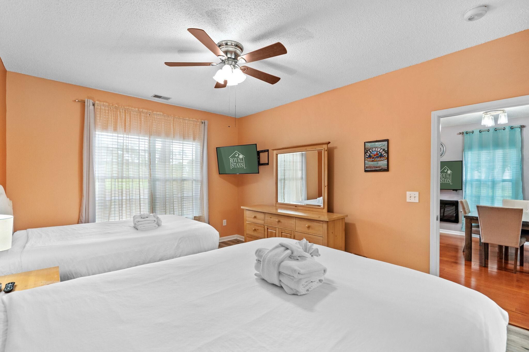 A hotel room with two beds, a ceiling fan, a wooden dresser, a mirror, and a dining area with a chair.