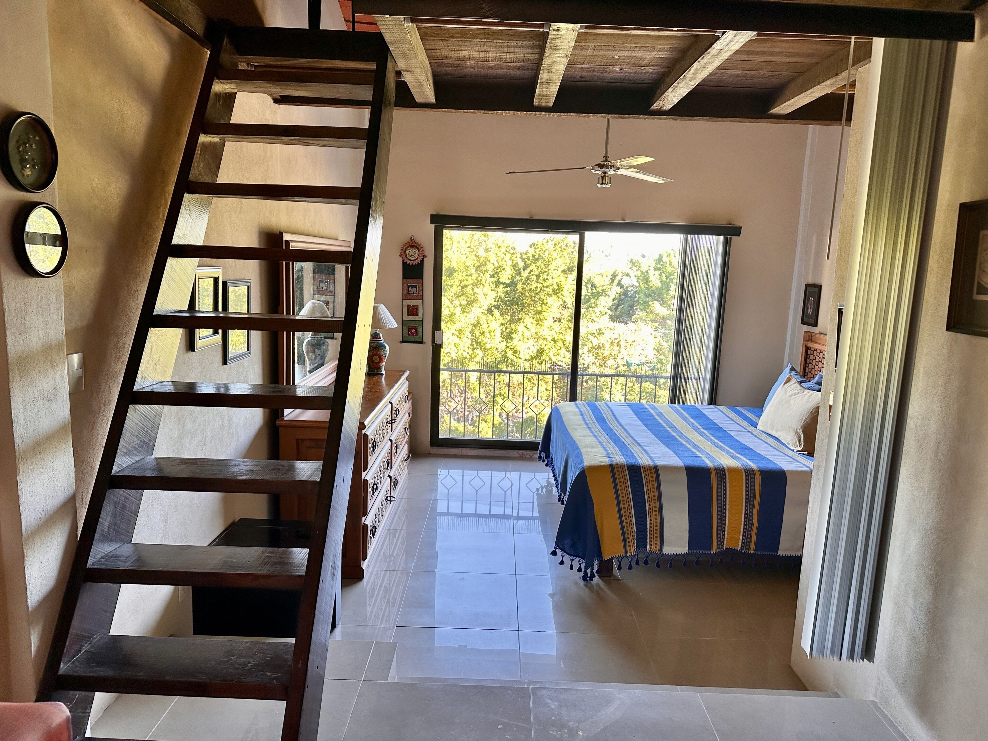 A bedroom with a bed, a ceiling fan, a window with a view, a staircase, and a door.