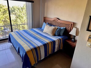 Ein Bett mit einem gemusterten Überwurf, ein Nachttisch mit einer Lampe und Blick auf Bäume durch ein Fenster.