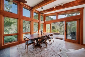 Ein geräumiges Zimmer mit einem großen Esstisch aus Holz, mehreren Stühlen und Blick auf eine Berglandschaft durch große Fenster.