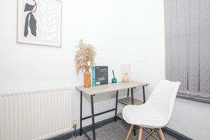 Ein Zimmer mit einem weißen Stuhl, einem Tisch mit einer Vase, einem gerahmten Bild und einer Heizung.