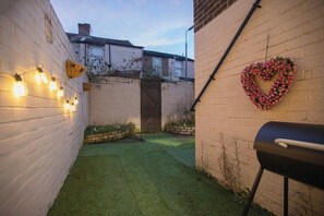 Une cour étroite, avec un mur de briques d’un côté, une porte en bois et, de l’autre, une décoration florale en forme de cœur.
