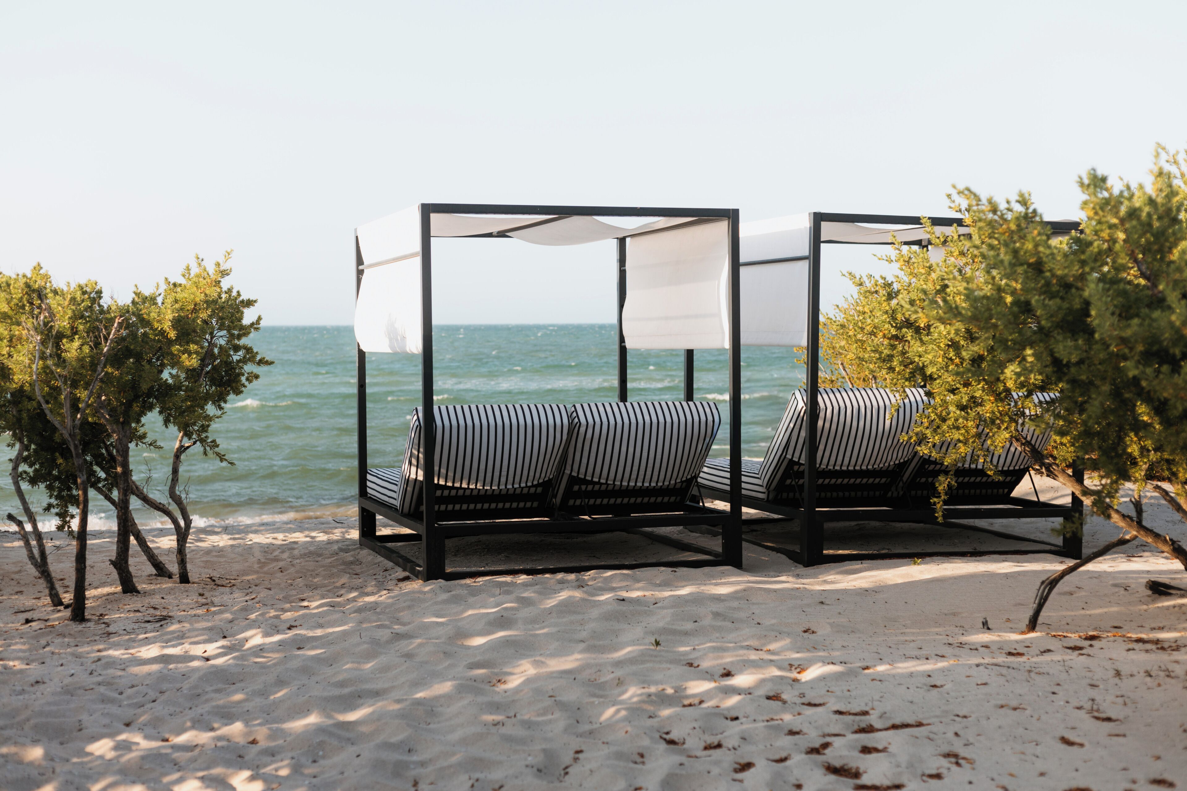 Ein Strandbett mit Baldachin, platziert am sandigen Ufer, mit Bäumen und dem Meer im Hintergrund.