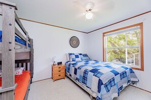 Un dormitorio con literas, una cama individual con colcha azul a cuadros, una mesita de noche con lámpara, y una ventana con vistas a árboles.