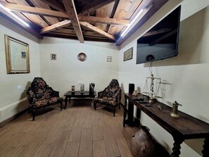 A room with wooden flooring, two ornate armchairs, a small table with decorative items, a television mounted on the wall, and a wooden cabinet with a brass object.