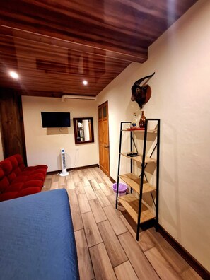 A bedroom with a bed, a red sofa, a wooden floor, a ceiling fan, a mounted TV, and a shelf with a decorative item.