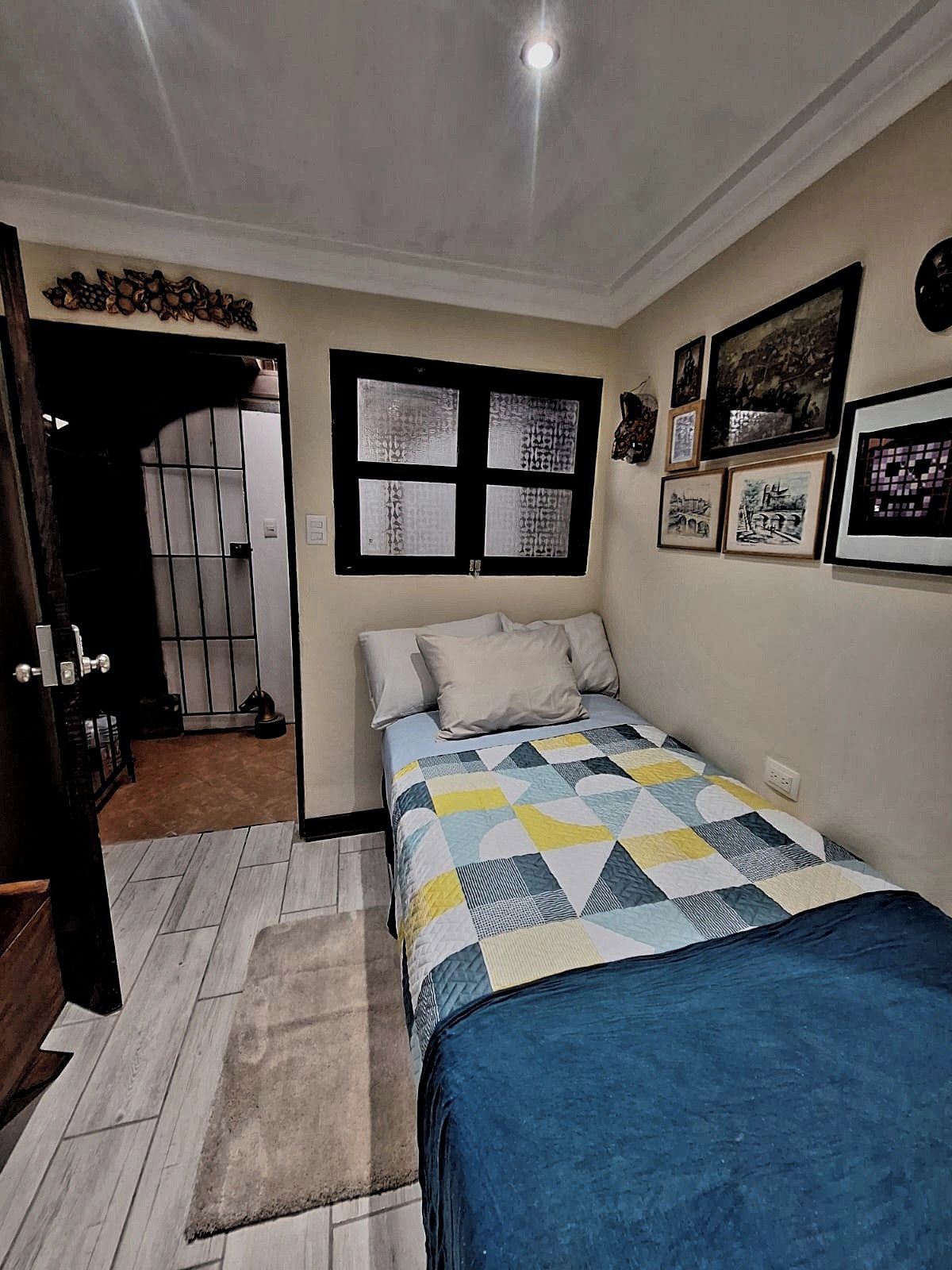 A bedroom with a bed, a window with blinds, a door, and a wall with framed pictures.