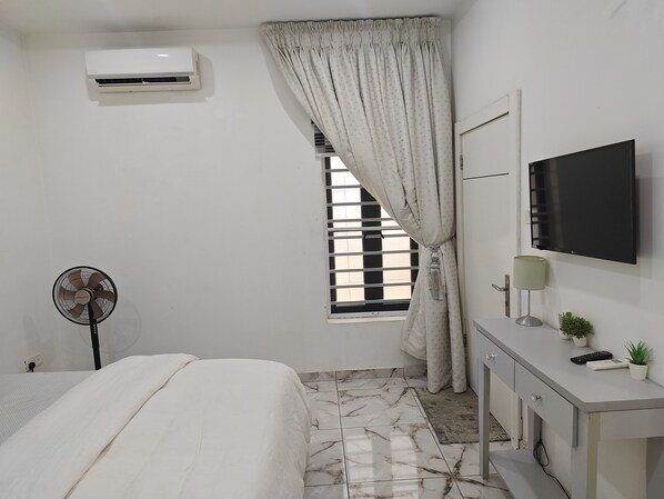 A bedroom with a bed, an air conditioner, a window with curtains, a television, a desk with a lamp, and a small plant.