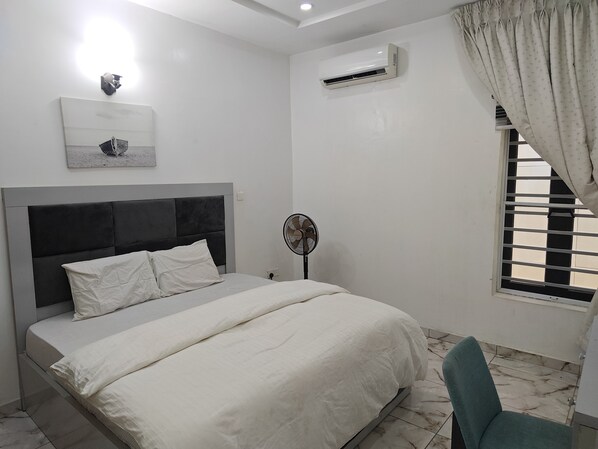 A bedroom with a bed, a standing fan, an air conditioning unit, and a window with blinds.