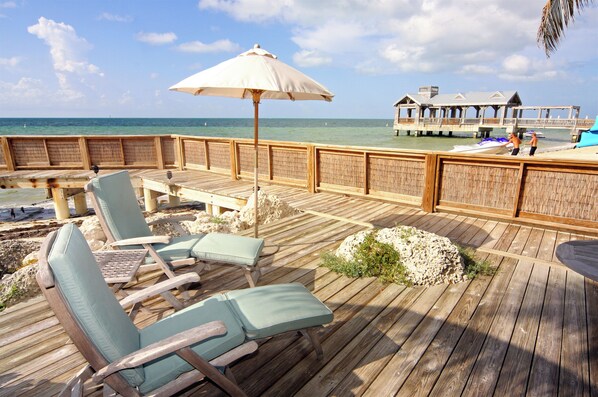 Eine Holzterrasse mit Liegestühlen und Sonnenschirm, mit Blick auf das Meer und einen Pier mit Gebäuden.