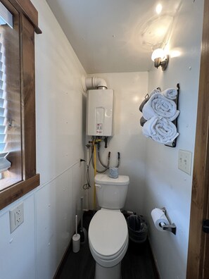 Ein kleines Badezimmer mit Toilette, einem weißen Tank, einem Handtuchhalter mit Handtüchern, einem Fenster mit Jalousien und einer Leuchte.