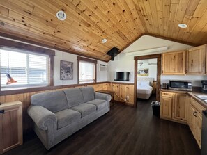 Ein gemütliches Interieur mit Holzbalkendecke, einem grauen Sofa, einer Küchenzeile und einem Fenster mit Jalousien.