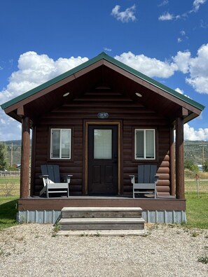 Eine Holzhütte mit einer dunkelbraunen Tür, je einem Fenster auf jeder Seite und einem grünen Dach. Vor der Veranda stehen zwei Adirondack-Stühle.