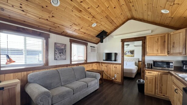 Un intérieur chaleureux avec un plafond en bois, un canapé gris, un coin cuisine et une fenêtre équipée de stores.