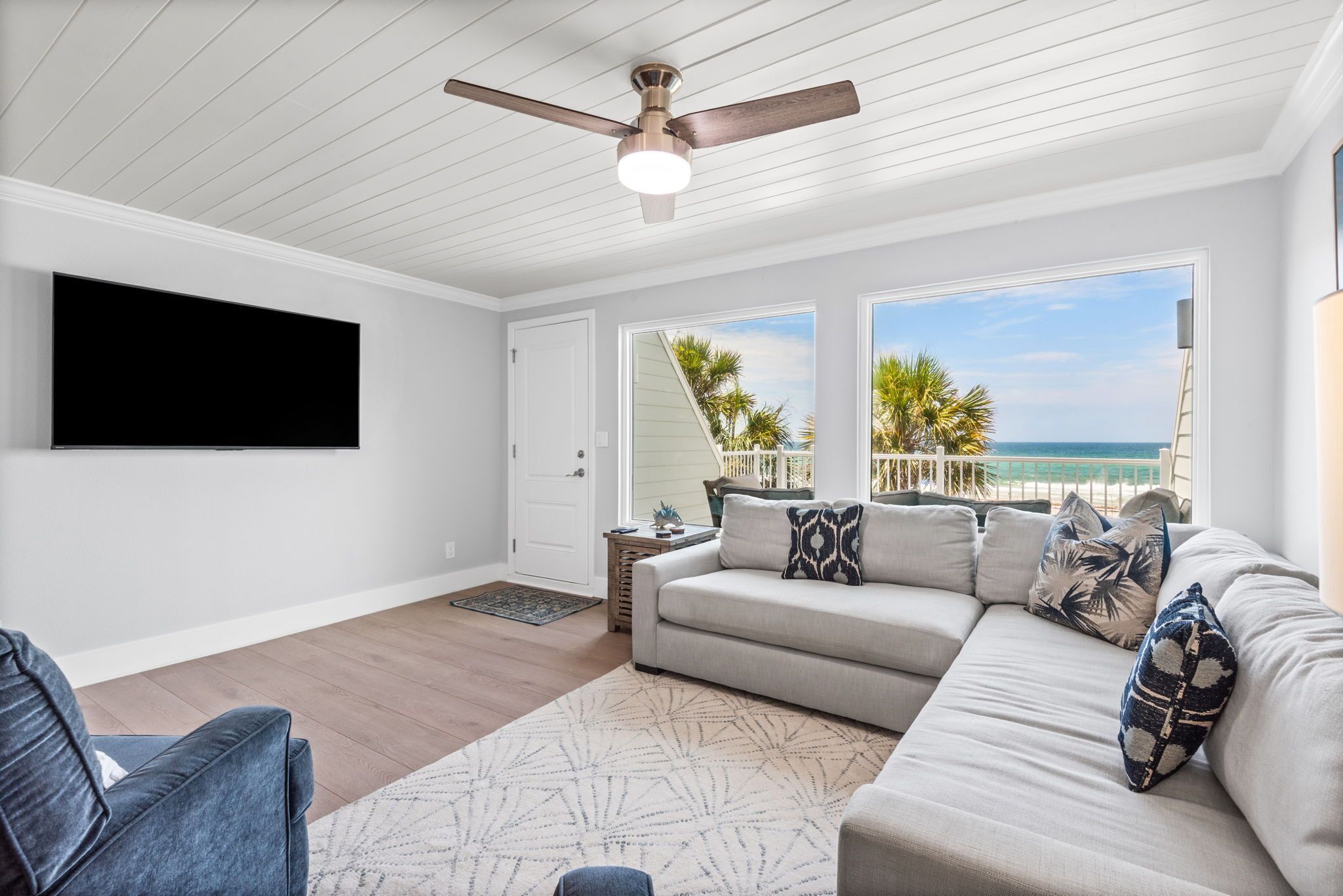 A living room with a ceiling fan, a large flat-screen TV, a sofa with patterned cushions, and a view of the ocean through large windows.