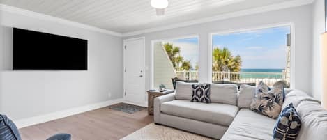 A living room with a ceiling fan, a large flat-screen TV, a sofa with patterned cushions, and a view of the ocean through large windows.