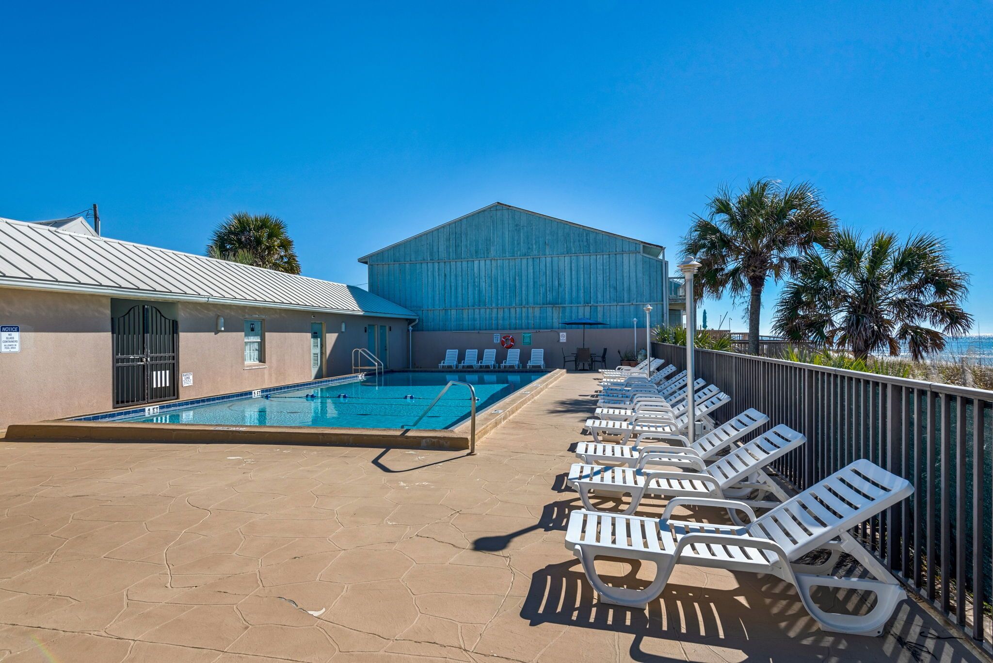 A pool area with lounge chairs, a building with a blue facade, and a clear view of the ocean.