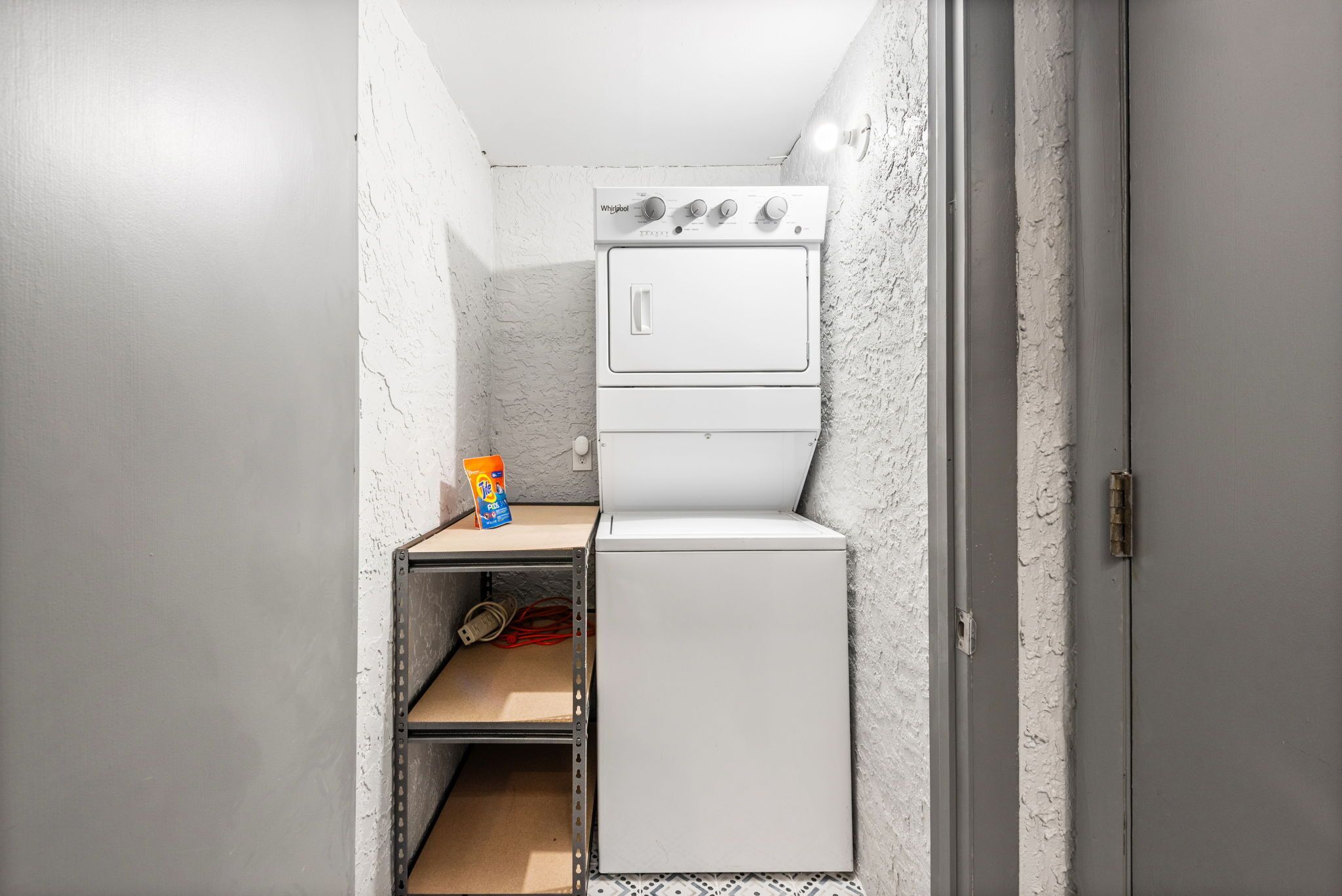 Une petite buanderie équipée d’un lave-linge et d’un sèche-linge superposés, d’une étagère sur laquelle se trouve un paquet de lessive, et d’une étagère métallique.