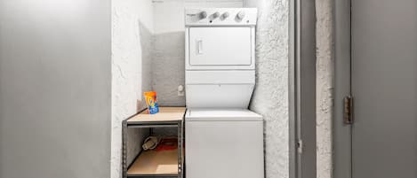 A compact laundry room with a stacked washer and dryer, a shelf with a bag of detergent, and a metal shelf unit.