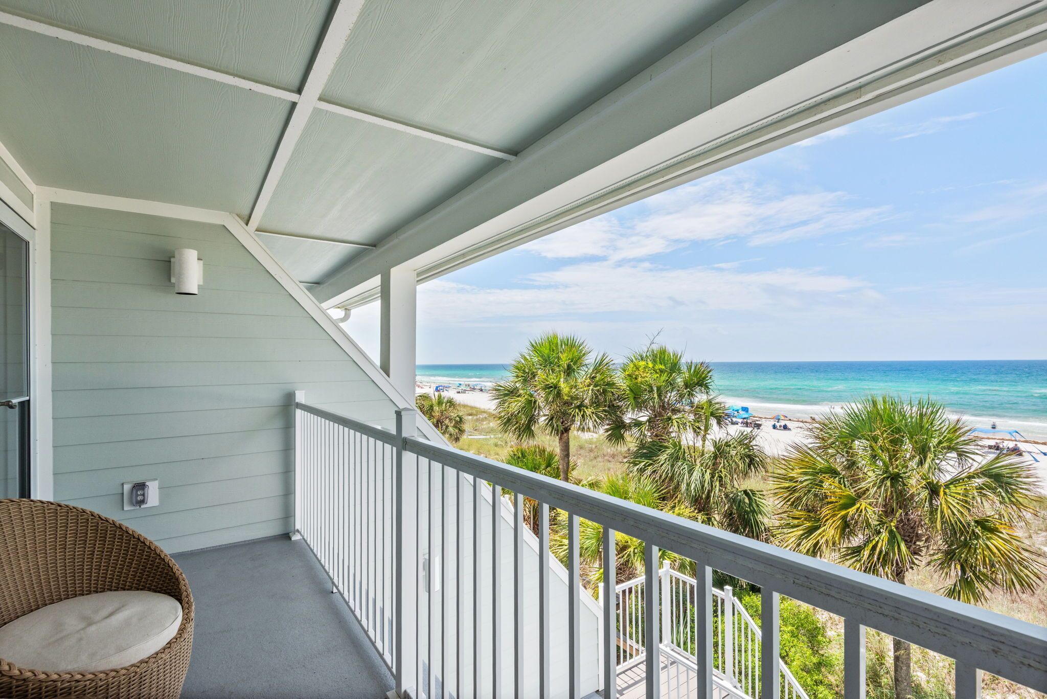 A balcony with a view of a beach, a wicker chair, and a white railing.