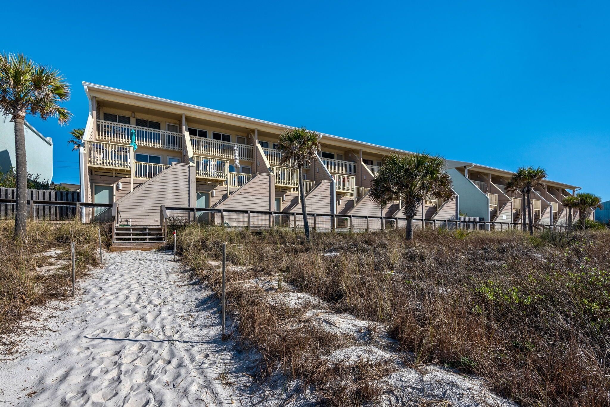 A beachfront hotel with a sandy path leading to the entrance, multiple balconies, and palm trees.