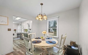 A dining area with a round table, white chairs, a chandelier, and a window with blinds.