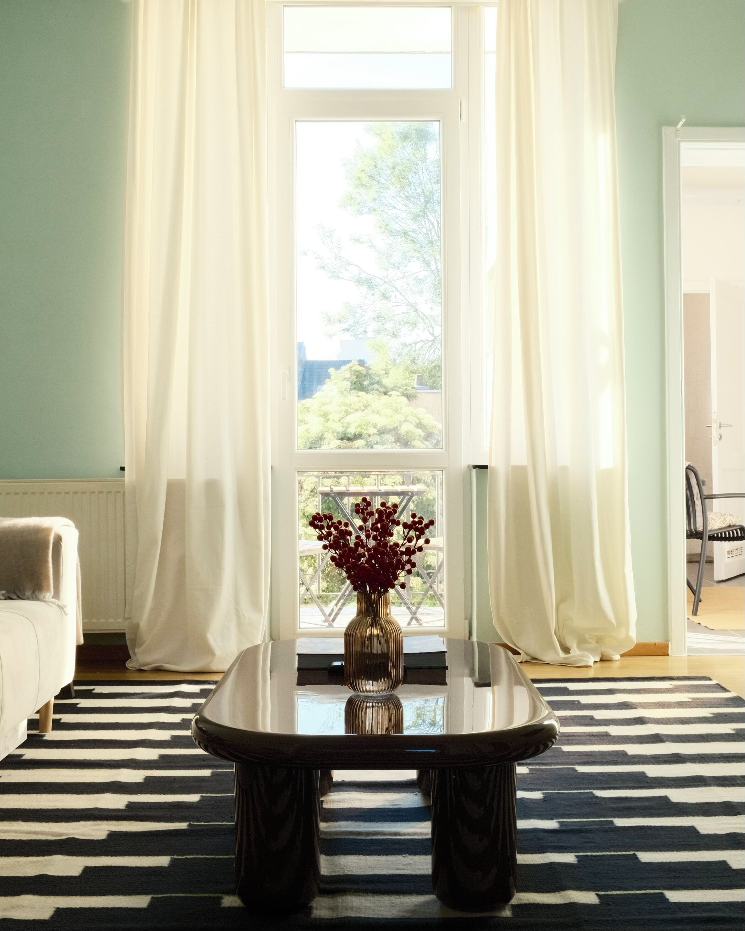 Une pièce avec une grande fenêtre, un tapis rayé noir et blanc et une table sur laquelle est posé un vase rempli de fleurs rouges.