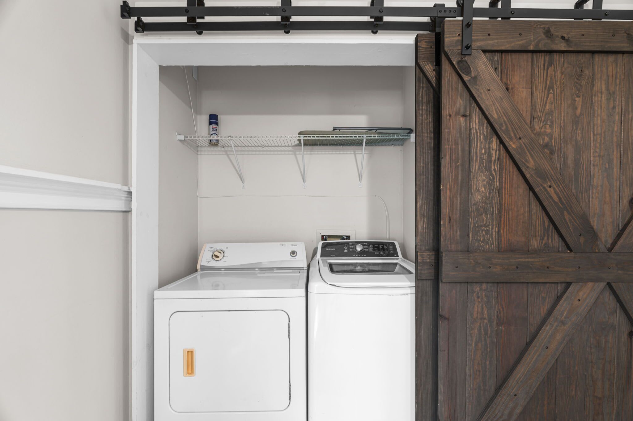 Une buanderie avec un lave-linge et un sèche-linge blancs, une étagère en métal sur laquelle se trouve une boîte de conserve, et une porte en bois de style grange.