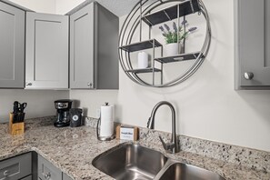 A kitchen with a granite countertop, stainless steel sink, and a round metal shelf with a potted plant. - Pier Escape A - WiFi, AC, Close to Beach (Jacksonville Beach)