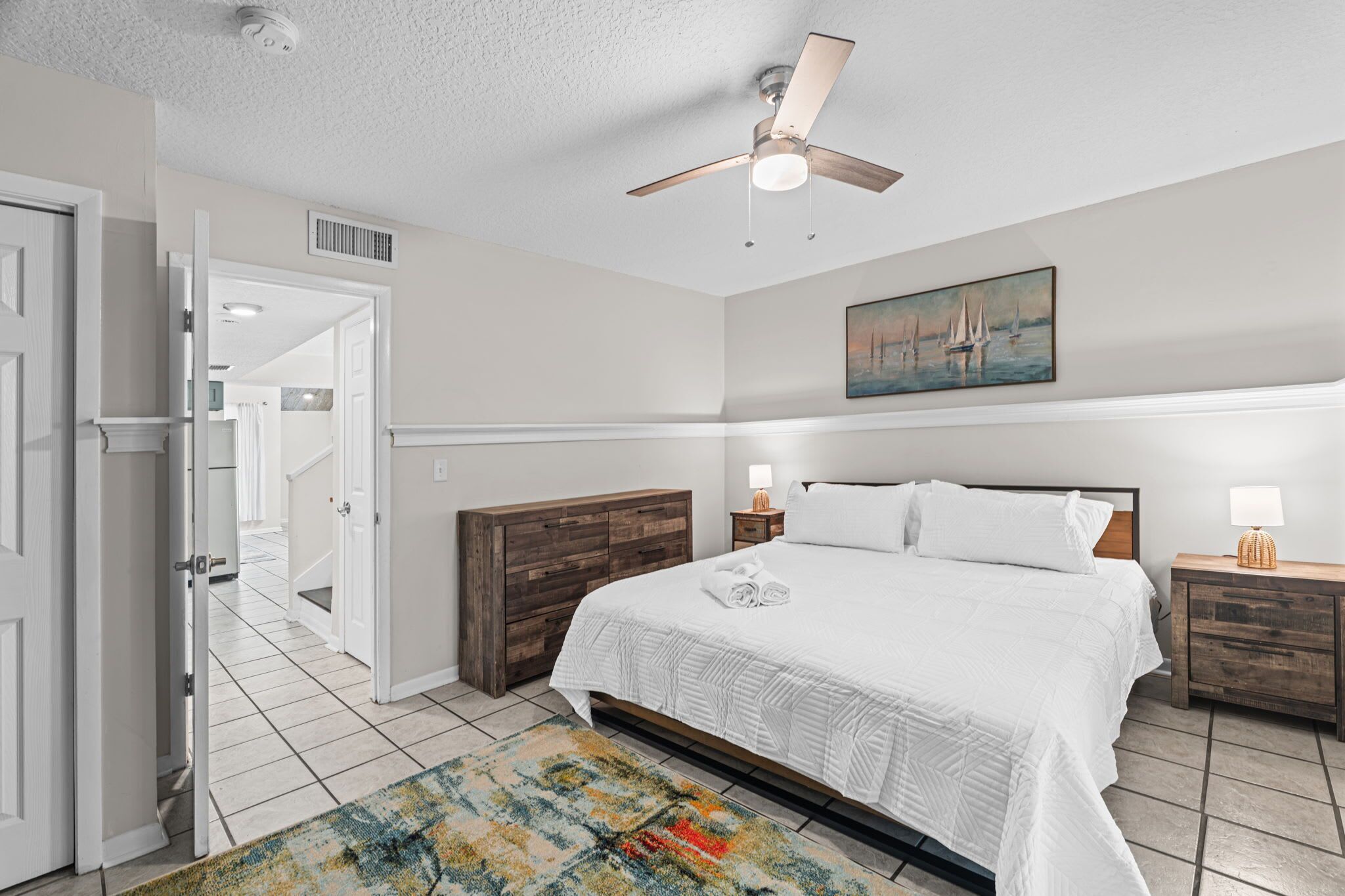 A bedroom with a bed, nightstands, a ceiling fan, a painting, and a door leading to another room.