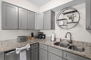 A modern kitchen with grey cabinets, a granite countertop, a coffee maker, a toaster, and a sink. - Pier Escape A - WiFi, AC, Close to Beach (Jacksonville Beach)