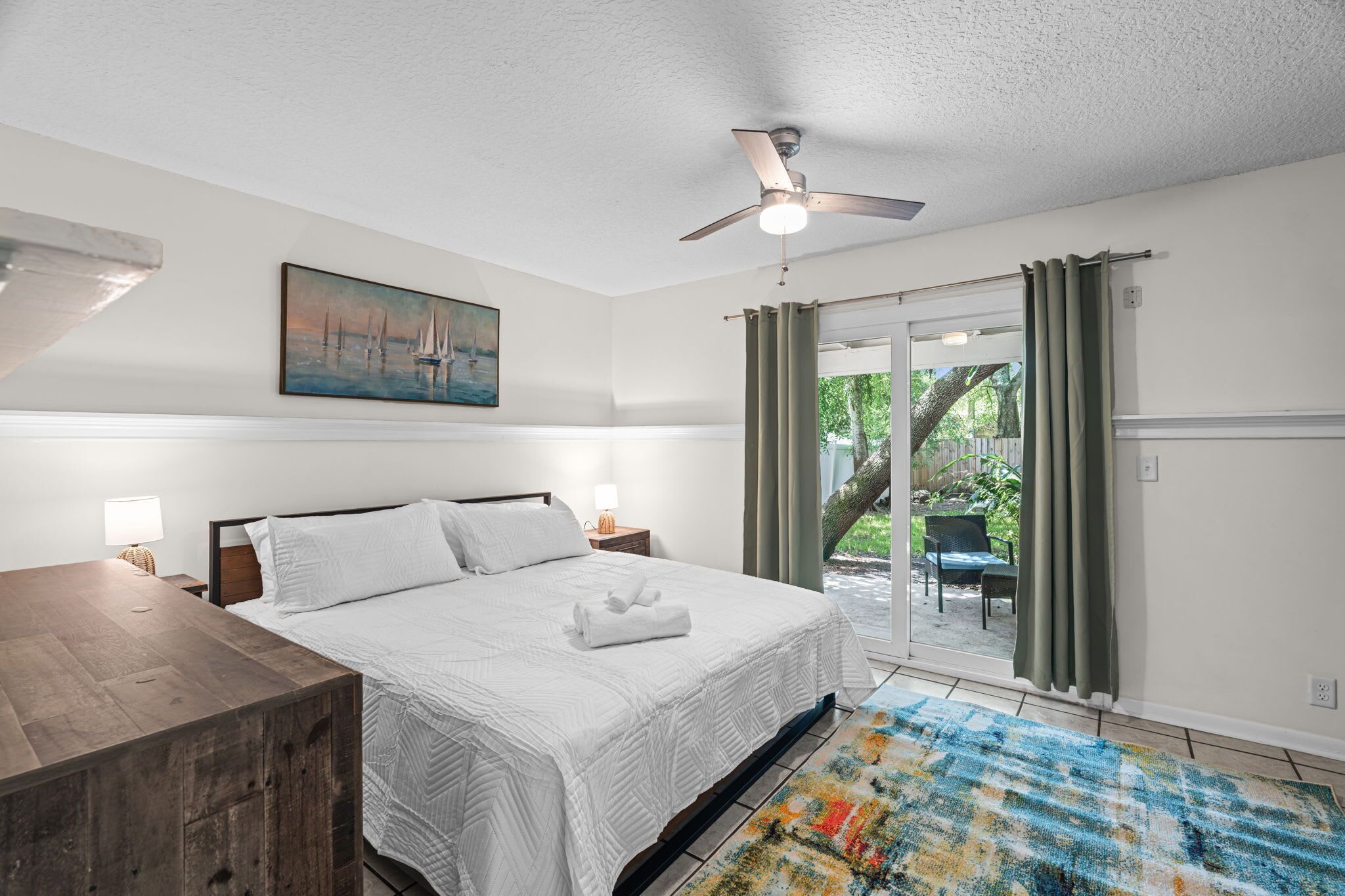 A bedroom with a bed, a painting, a ceiling fan, and a view of the outdoors.
