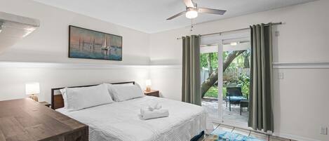 A bedroom with a bed, a painting, a ceiling fan, and a view of the outdoors.