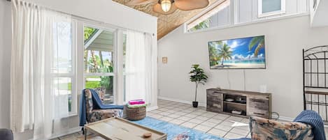 A spacious living room with a ceiling fan, a large TV, and a view of the outdoors through a sliding glass door.