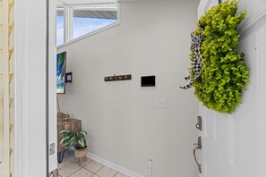 A hallway with a wooden door, a small table with a plant, a wall-mounted shelf with decorative items, and a window with a view of the sky. - Pier Escape - Entire Property (Jacksonville Beach)