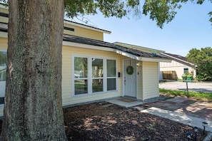 A single-story residential building with a front door, large windows, and a tree in the foreground. - Pier Escape B - WiFi, AC, Close to Beach (Jacksonville Beach)
