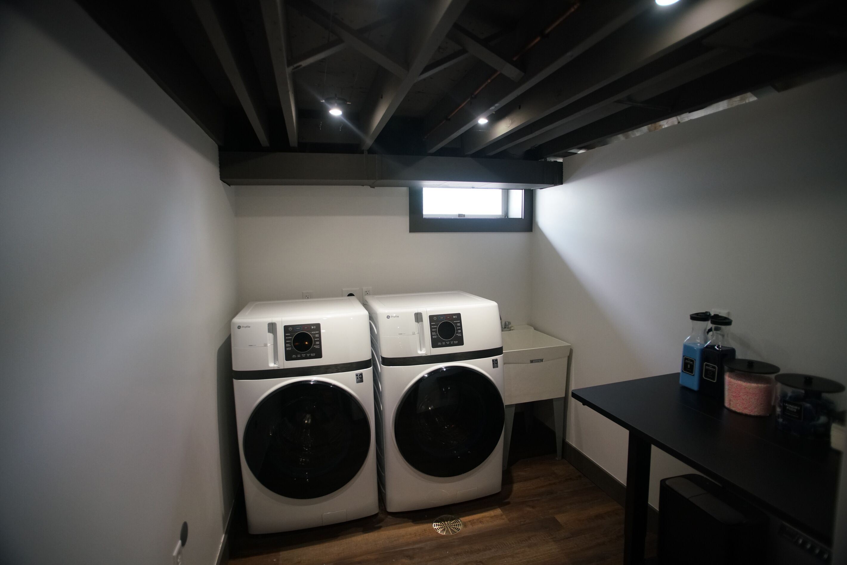 A laundry room with two front-loading washing machines, a small window, and a table with cleaning supplies.