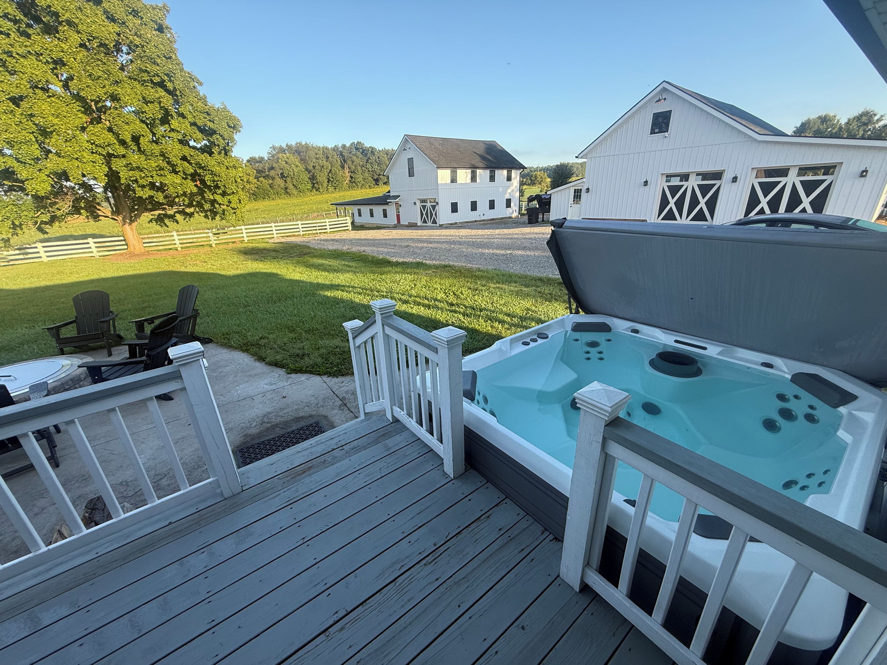 A deck with a hot tub, chairs, and a view of a barn and trees.