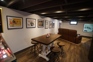 A room with a wooden table, stools, a cushioned bench, and framed movie posters on the wall. - Black, White, & Barn w/ Hot Tub & Fire Pit (Fredericktown)