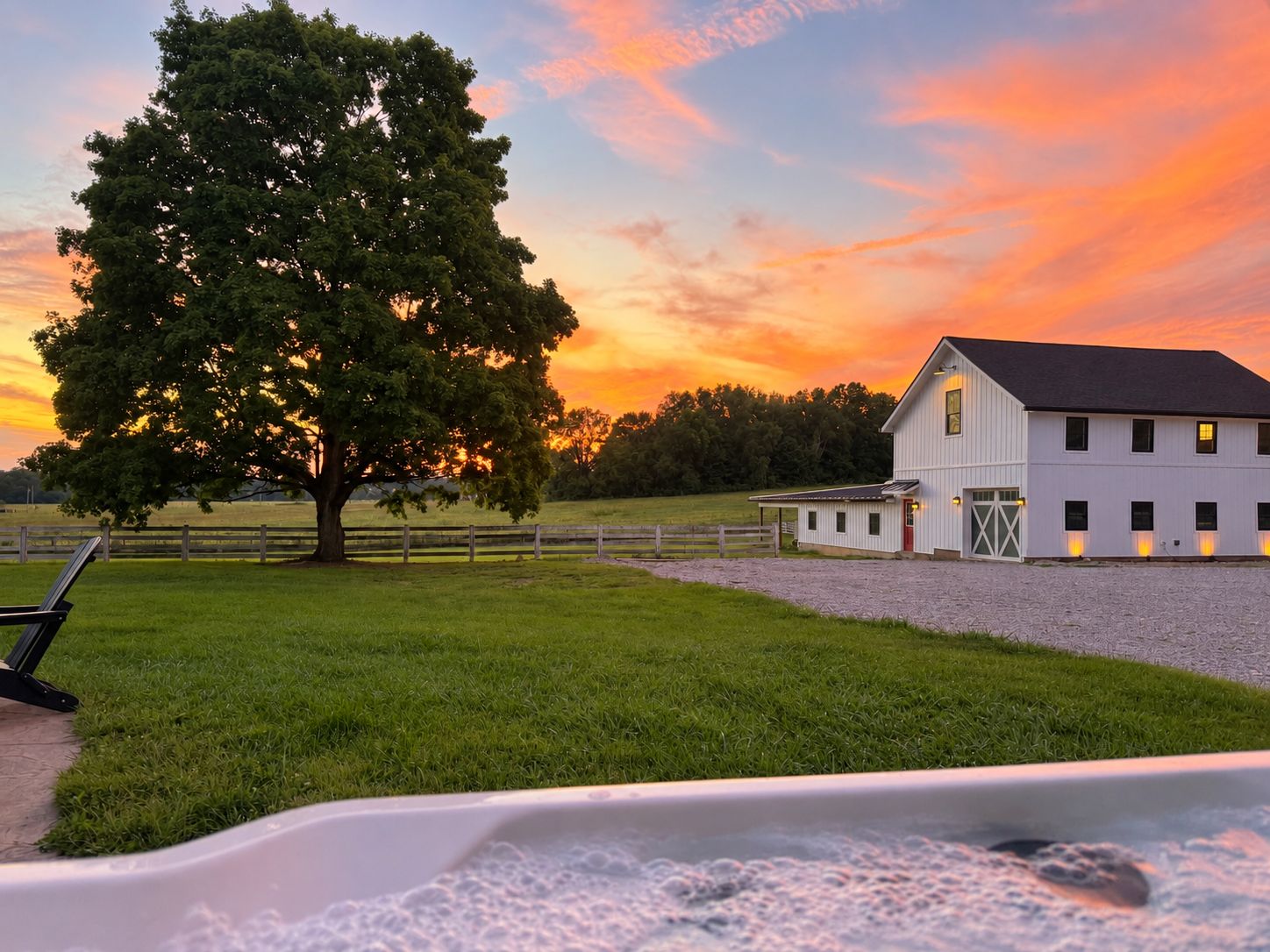 A barn with a large tree in the foreground, a fenced area, and a clear sky.