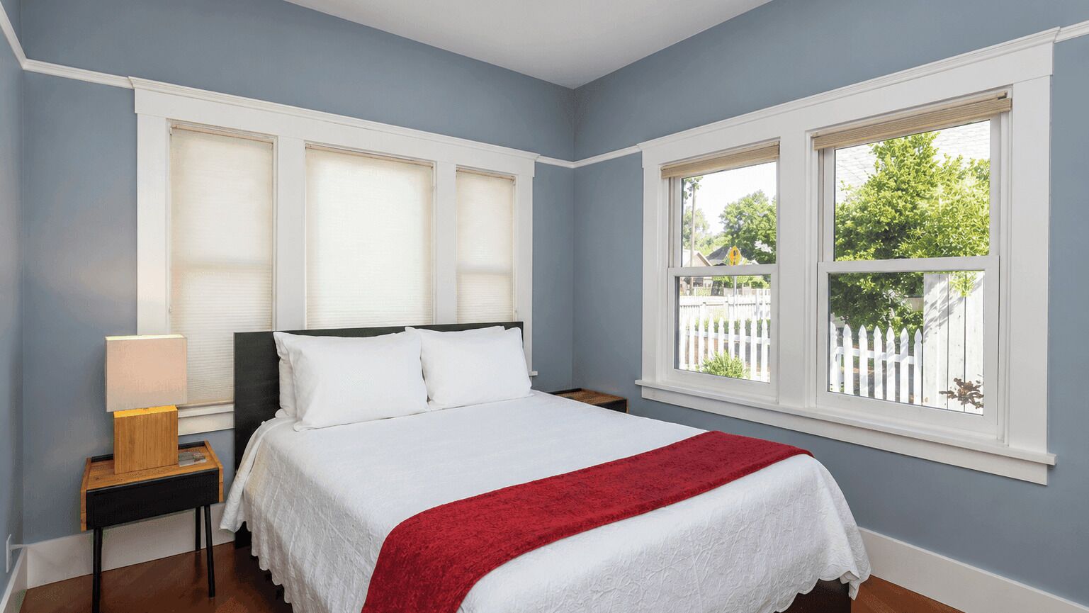 A bedroom with a bed, a nightstand, a lamp, and a window with a view of trees and a white fence.