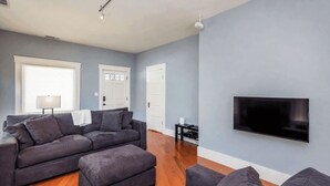 Ein Wohnzimmer mit grauem Sofa, einem flachbildfernseher an der Wand und einer Lampe auf einem Beistelltisch.