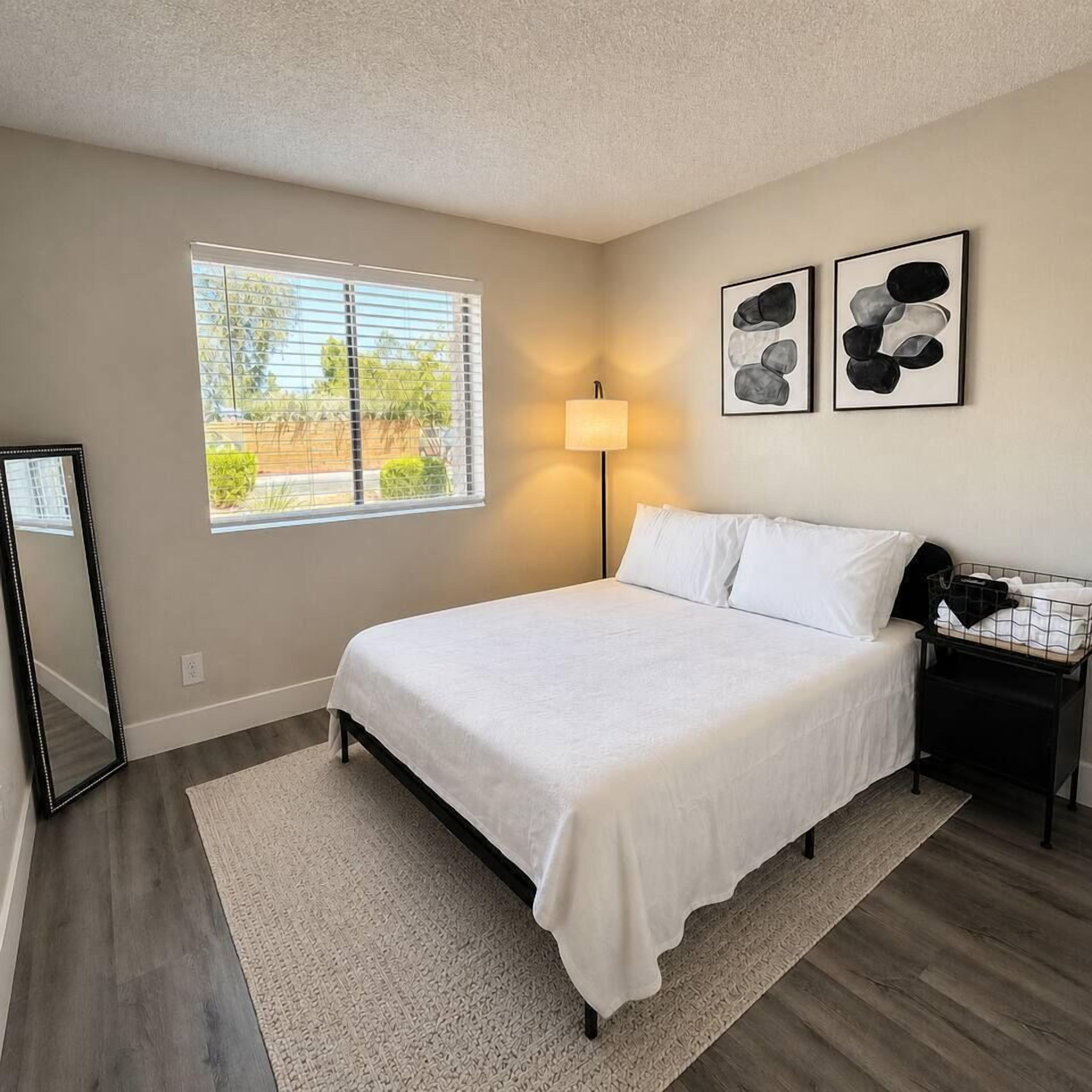 A bedroom with a bed, a bedside table, a lamp, a window with blinds, and two framed pictures on the wall.