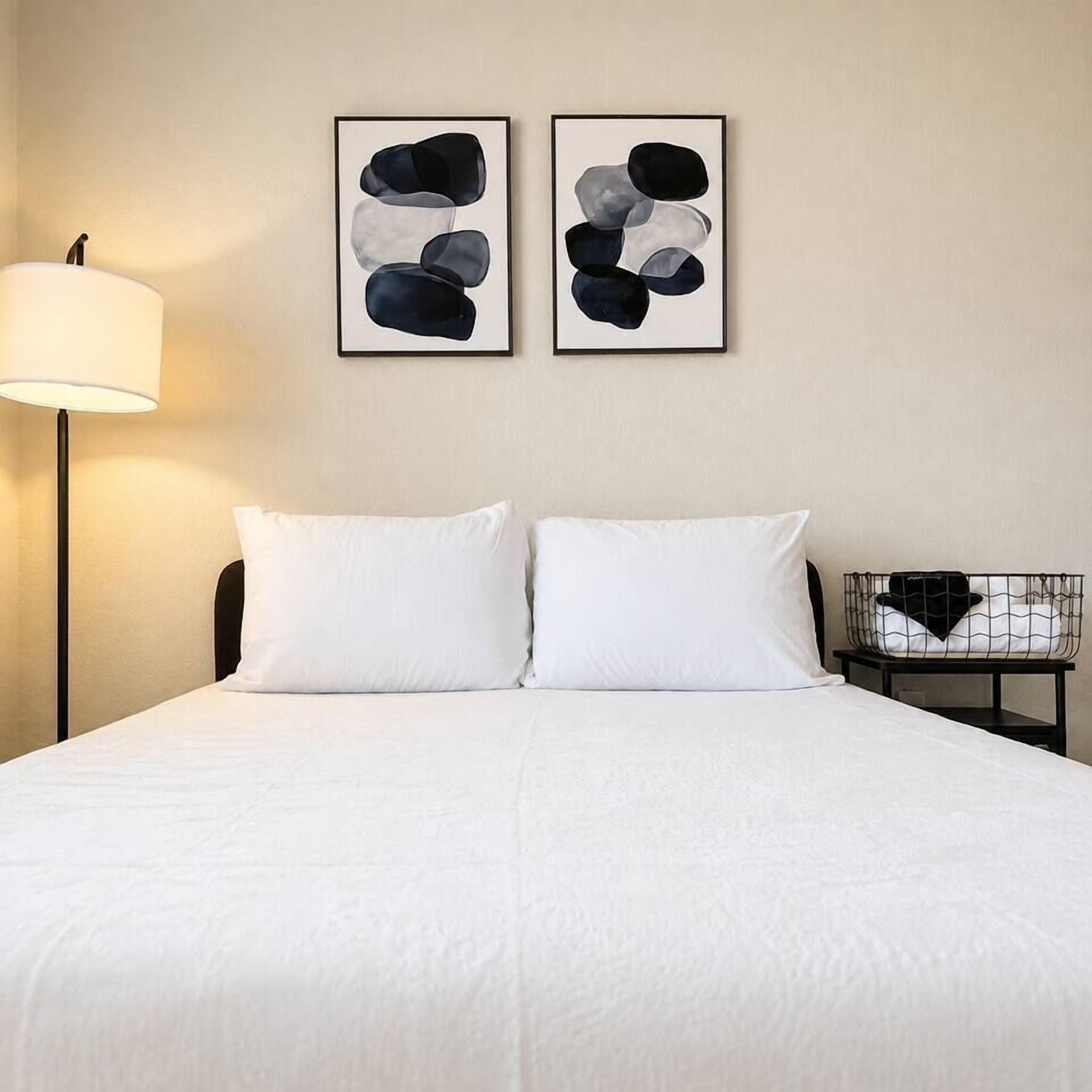 A hotel room with a bed, two pillows, a nightstand with a lamp, and two abstract paintings on the wall.