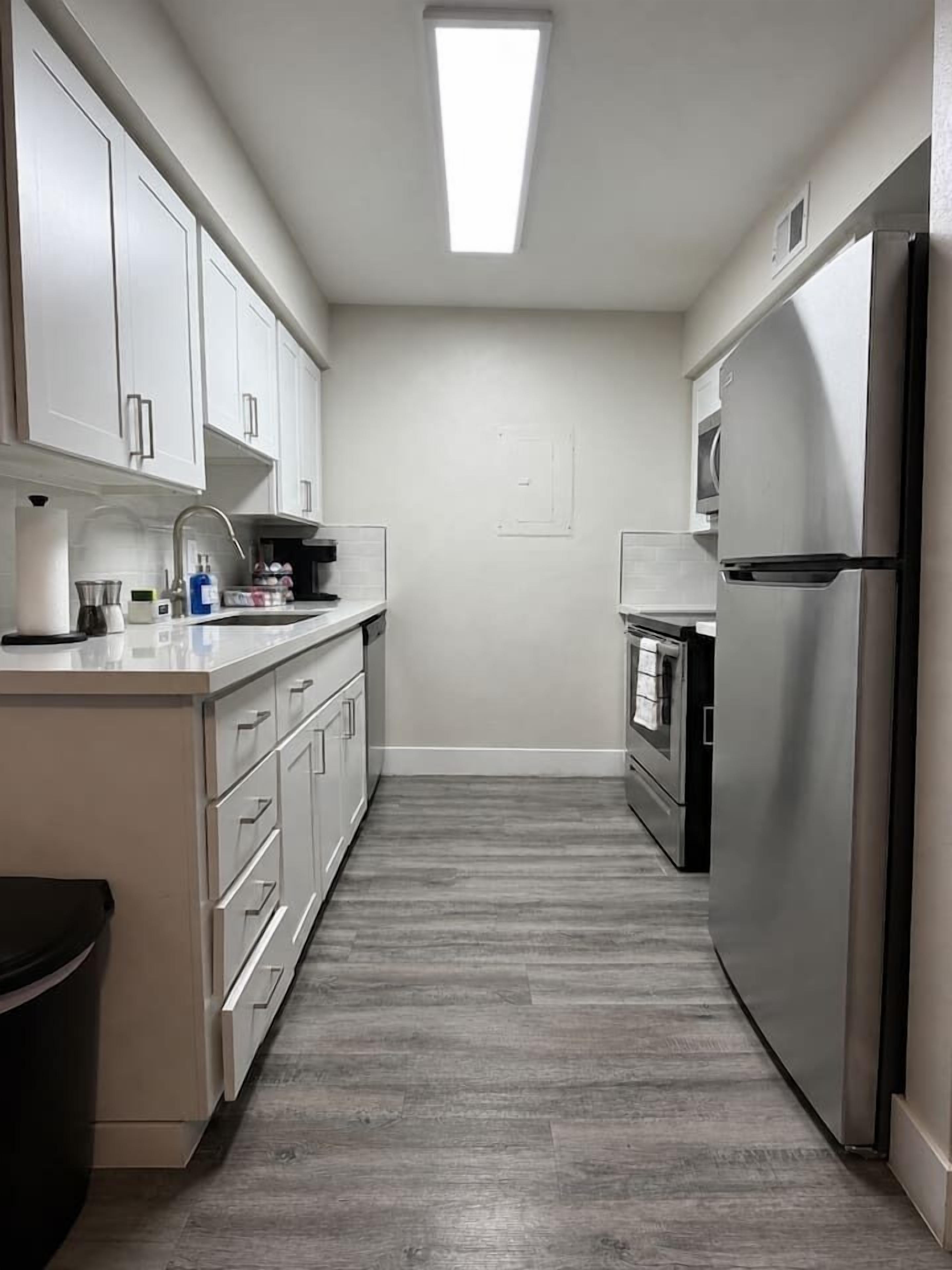 A narrow kitchen with white cabinets, a stainless steel refrigerator, and a built-in oven.