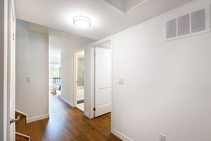 A hallway with wooden flooring, white walls, and a door leading to a bathroom with a bathtub.