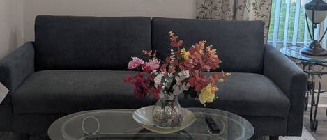 A living room with a sofa, a glass coffee table with a vase of flowers, a framed picture on the wall, and a window with curtains.