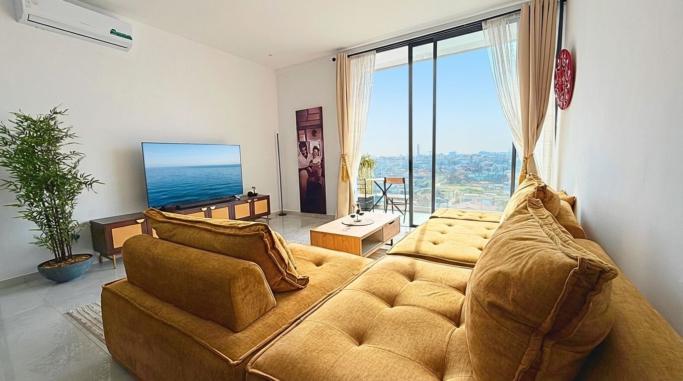 Un salon moderne avec un canapé, un téléviseur à écran plat, une plante en pot et une vue sur la ville grâce à de grandes fenêtres.